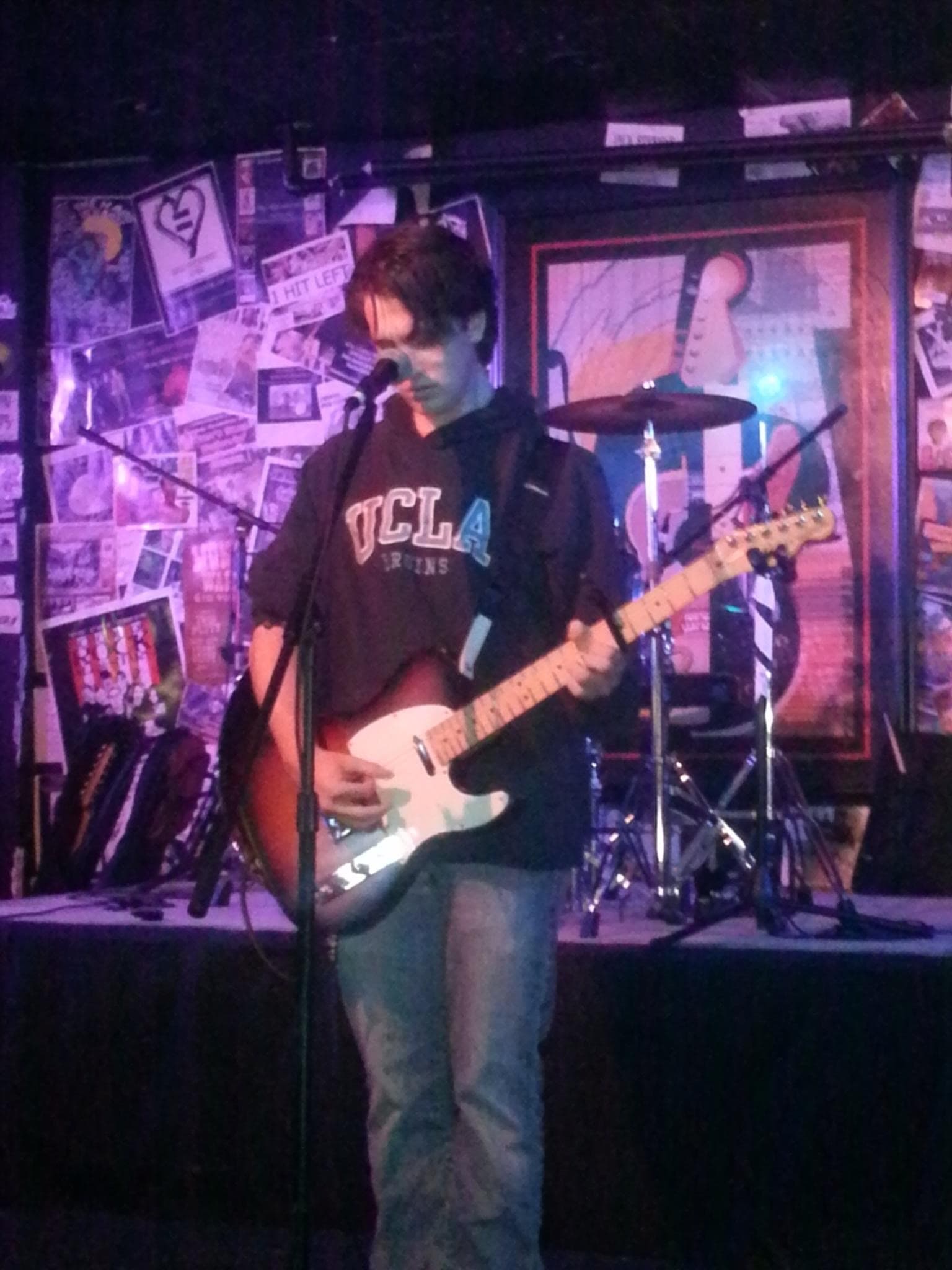 Johnny Suede on stage with a Telecaster, UCLA hoodie, mic stand and drum kit visible behind