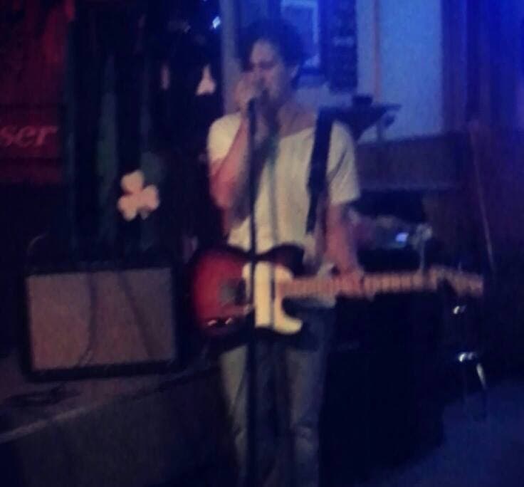 Johnny Suede at the mic, white tee, holding a Telecaster, amp and stage lighting in frame